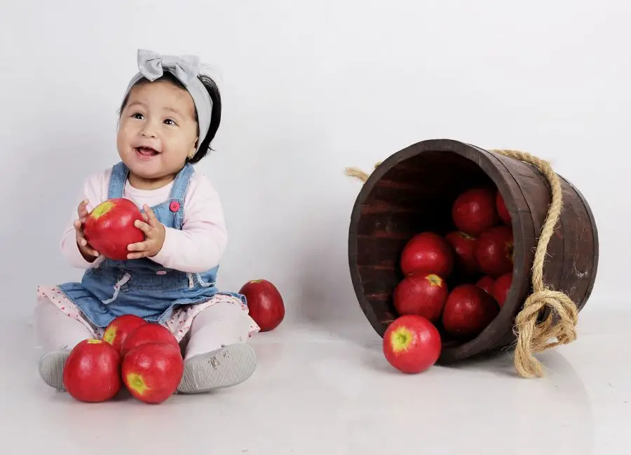 Dziewczynka siedząca obok przewróconego, drewnianego wiaderka trzyma w rączkach jabłko.